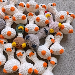 Op de afbeelding: Een verzameling handgemaakte gehaakte ganzen-speeltjes. De ganzen zijn voornamelijk wit met oranje accenten op hun snavels en voeten. Sommige ganzen hebben verschillende kleuren, waaronder roze, groen en bruin. De speeltjes liggen op een grijze ondergrond.