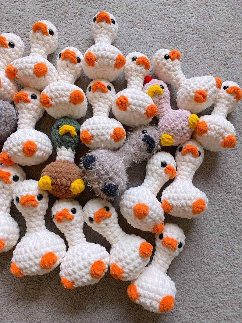 Op de afbeelding: Een verzameling handgemaakte gehaakte ganzen-speeltjes. De ganzen zijn voornamelijk wit met oranje accenten op hun snavels en voeten. Sommige ganzen hebben verschillende kleuren, waaronder roze, groen en bruin. De speeltjes liggen op een grijze ondergrond.