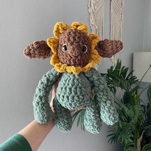 May include: A handmade crochet sunflower plush toy. The toy features a brown head with a yellow and brown sunflower design, brown ears, and green body and limbs. The plush is held up in front of a gray wall.