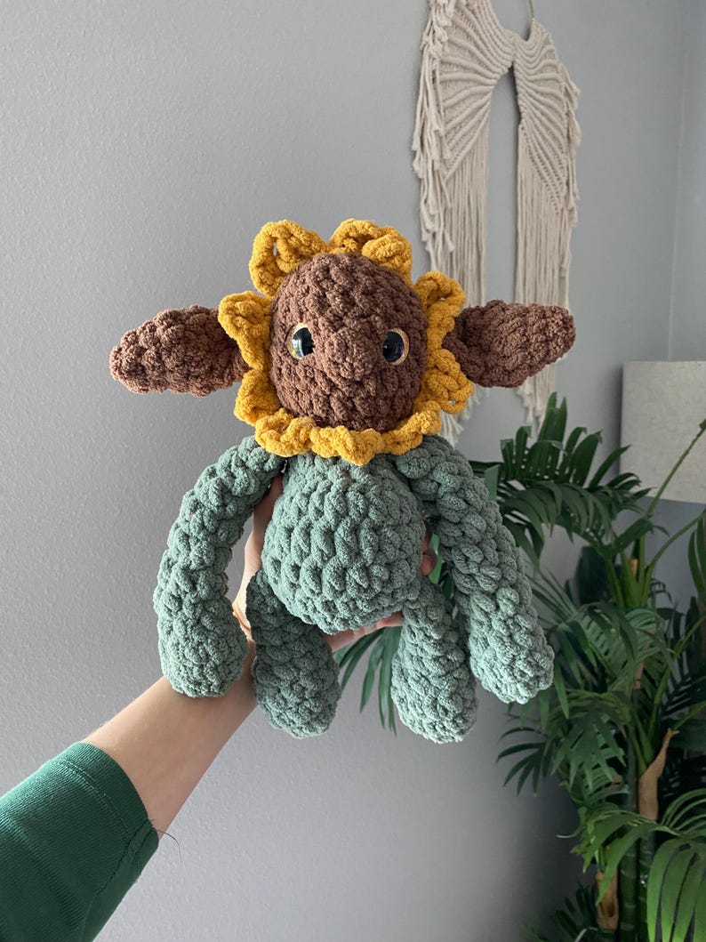 May include: A handmade crochet sunflower plush toy. The toy features a brown head with a yellow and brown sunflower design, brown ears, and green body and limbs. The plush is held up in front of a gray wall.