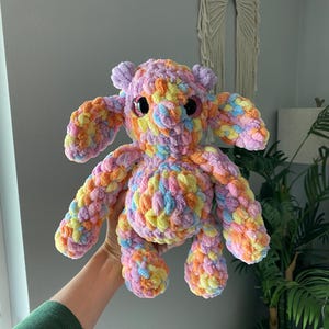 May include: A handmade, colorful crochet creature with large, round eyes. The plush toy features a mix of pastel colors, including pink, yellow, orange, and blue. It has large ears and stubby limbs, giving it a whimsical appearance.