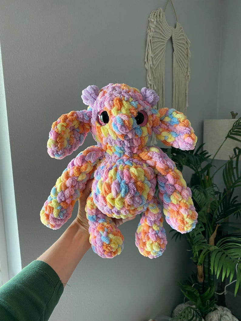 May include: A handmade, colorful crochet creature with large, round eyes. The plush toy features a mix of pastel colors, including pink, yellow, orange, and blue. It has large ears and stubby limbs, giving it a whimsical appearance.