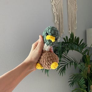 Op de afbeelding: Een handgemaakte gehaakte eend met een groene kop, gele snavel, bruin lichaam en gele voeten. De eend wordt in een hand gehouden, wat de zachte textuur en gedetailleerde afwerking laat zien. Dit schattige pluche speelgoed is perfect voor kinderkamers of als uniek cadeau.