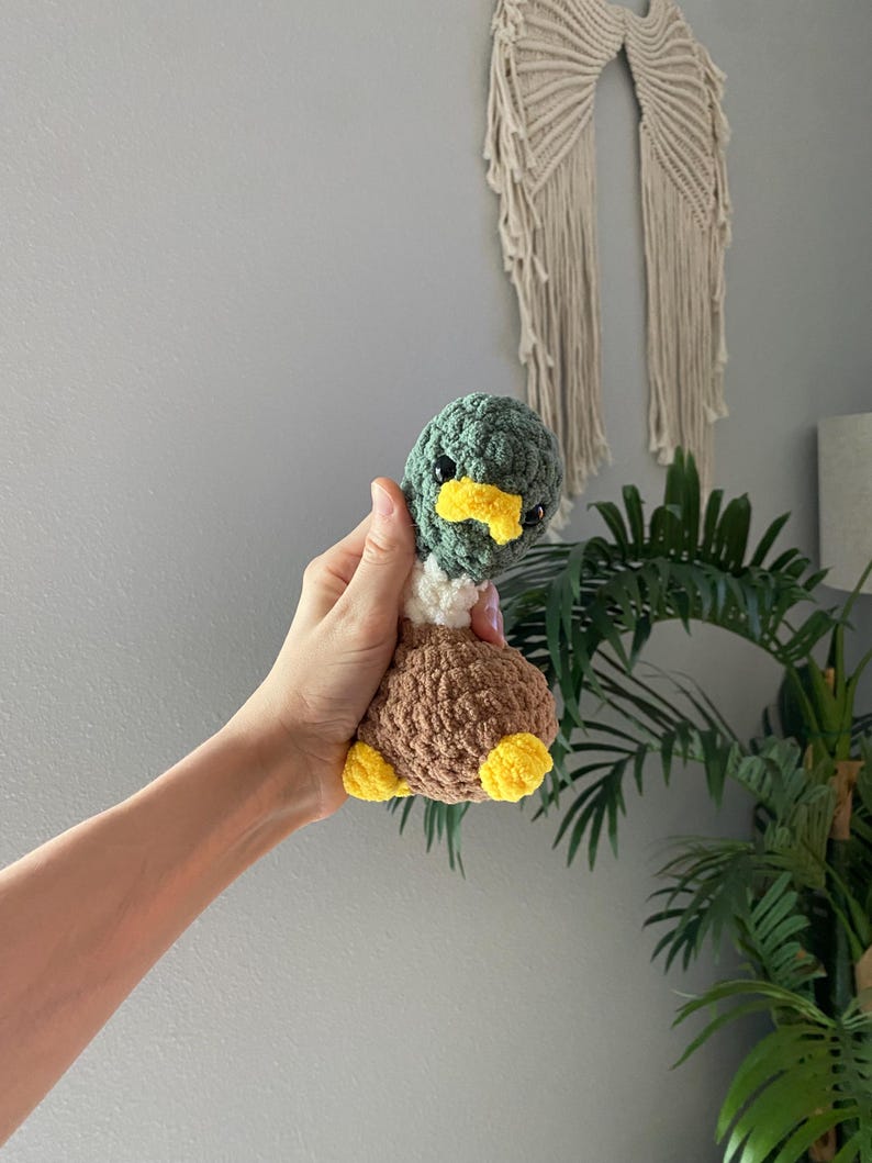 Op de afbeelding: Een handgemaakte gehaakte eend met een groene kop, gele snavel, bruin lichaam en gele voeten. De eend wordt in een hand gehouden, wat de zachte textuur en gedetailleerde afwerking laat zien. Dit schattige pluche speelgoed is perfect voor kinderkamers of als uniek cadeau.