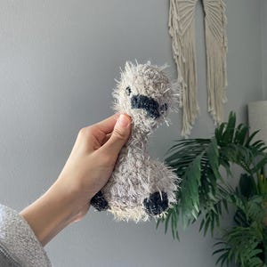 Op de afbeelding: Een handgemaakt, grijs en zwart gehaakt lama pluche speelgoed. De lama heeft een harige textuur, zwarte accenten op zijn gezicht en voeten en kleine zwarte ogen. Het pluche wordt in de hand van een persoon gehouden.