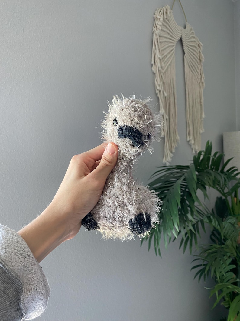 Op de afbeelding: Een handgemaakt, grijs en zwart gehaakt lama pluche speelgoed. De lama heeft een harige textuur, zwarte accenten op zijn gezicht en voeten en kleine zwarte ogen. Het pluche wordt in de hand van een persoon gehouden.