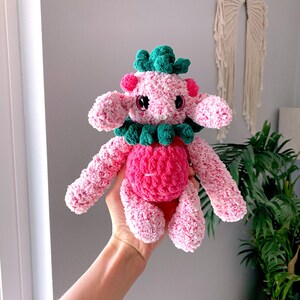 May include: A handmade pink and green crochet plush toy. The plush has a round pink body, pink arms and legs, and a green collar and headpiece. The toy has black eyes and is held in a person's hand.