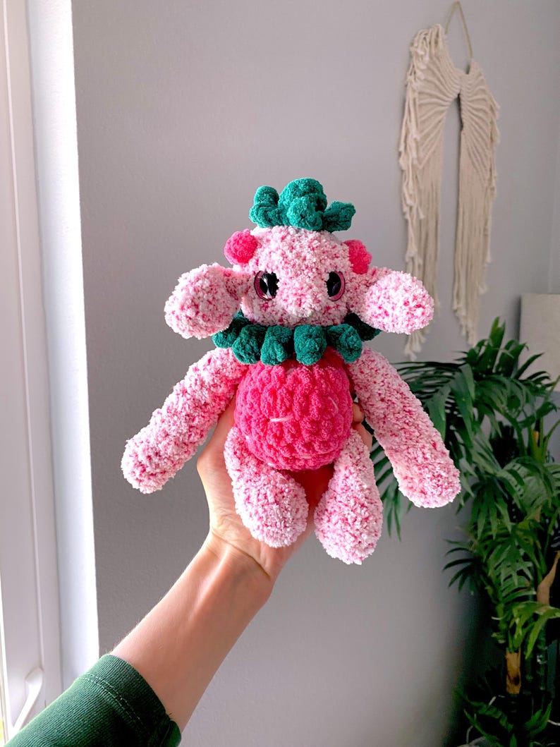 May include: A handmade pink and green crochet plush toy. The plush has a round pink body, pink arms and legs, and a green collar and headpiece. The toy has black eyes and is held in a person's hand.