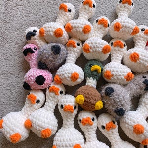 Op de afbeelding: Een verzameling handgemaakte gehaakte ganzen in verschillende kleuren. De meeste zijn wit met oranje voeten en accenten, terwijl andere roze, grijs en bruin zijn. Elke gans heeft zwarte ogen en een oranje snavel.