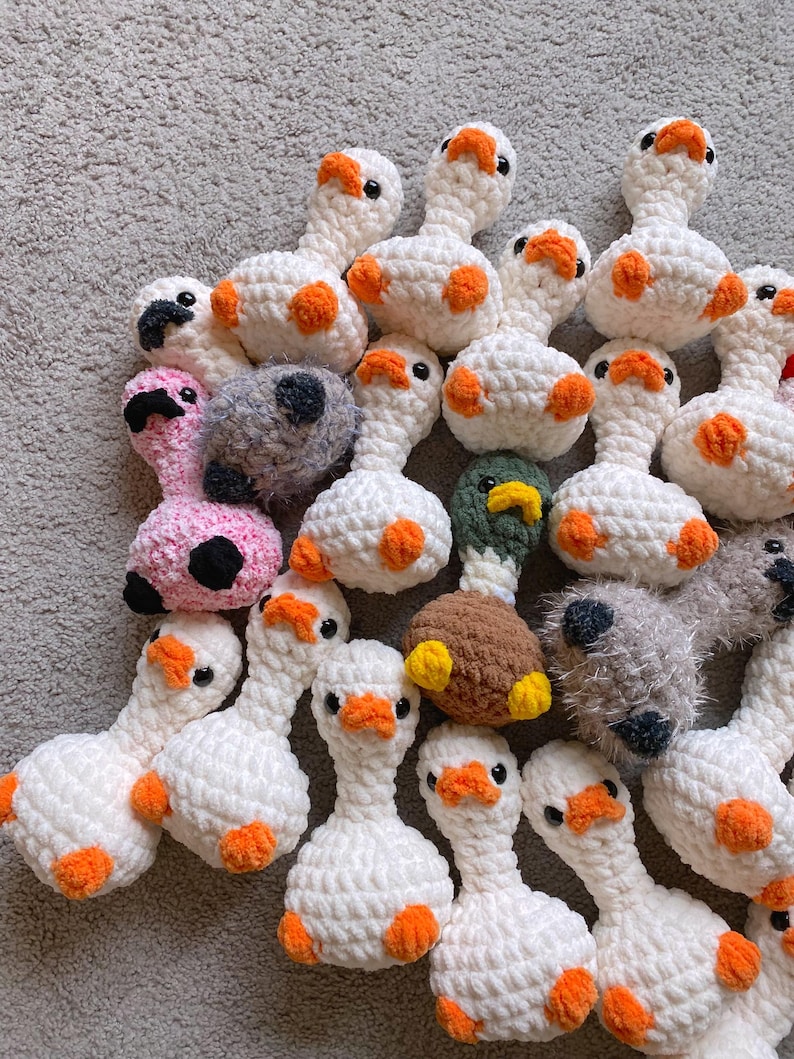 Op de afbeelding: Een verzameling handgemaakte gehaakte ganzen in verschillende kleuren. De meeste zijn wit met oranje voeten en accenten, terwijl andere roze, grijs en bruin zijn. Elke gans heeft zwarte ogen en een oranje snavel.
