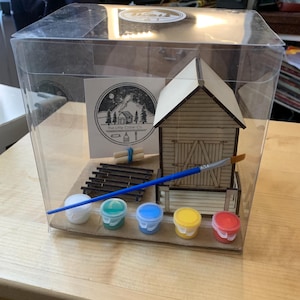 May include: A miniature wooden barn kit presented in a clear plastic box. The kit contains a small wooden barn, paint pots in white, green, blue, yellow, and red, a paintbrush, and other accessories. The kit is from "The Little Cabin Co."
