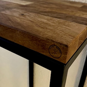 Könnte beinhalten: Eine Tischplatte aus Holz mit einem schwarzen Metallrahmen. Die Tischplatte hat eine natürliche Holzmaserung und einen schwarzen Stempel mit der Aufschrift "PATINE DESIGN" mit einem Kreuz und dem Jahr "EST. 2000".