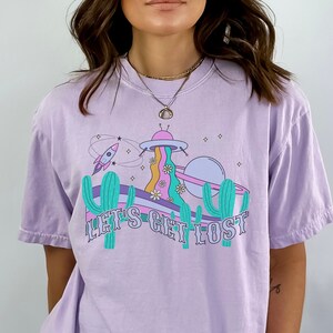 May include: A lilac t-shirt with a graphic design featuring a rocket ship, cactus, and a planet with the text "LET'S GET LOST". The design has a retro, whimsical style with pastel colours.