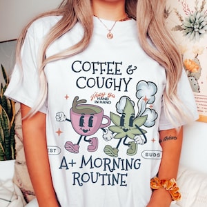 May include: White t-shirt with a graphic that reads "Coffee & Coughy" and "A+ Morning Routine." The design features cartoon coffee and cannabis characters. The shirt is a casual, everyday wear item.