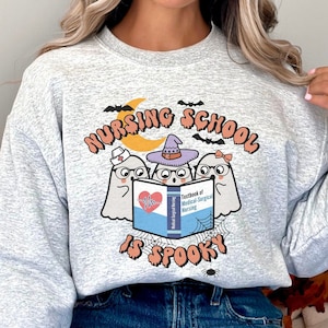 May include: A gray sweatshirt with the text "Nursing School is Spooky" and a graphic of three ghosts reading a book. The ghosts are wearing hats and glasses.