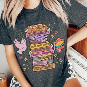 May include: A dark grey heather t-shirt with a colorful graphic of a stack of books. The books have text on them that reads "Naloxone", "Needle Exchange Programs", "MAT Programs", "Safe Use Sites", "Education", "Multiple Pathways", "Outreach Programs", "Peer Support", "Acceptance". There is a pink bird and a butterfly on the graphic.