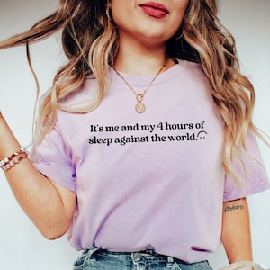 May include: A woman wearing a light purple t-shirt with the text "It's me and my 4 hours of sleep against the world." The shirt is a relaxed fit and has a crew neck. She is wearing blue jeans and a brown and white plaid scarf.