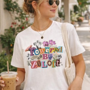 May include: White t-shirt with the text "Powered By Zoloft" in colorful block letters and floral accents. The person is wearing sunglasses and carrying a tote bag and a beverage in a clear plastic cup with a straw.