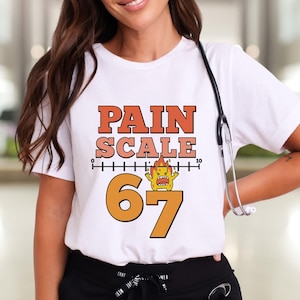 May include: White t-shirt with the words "PAIN SCALE" in orange and a pain scale graphic. Below the text is the number "67" in orange. A cartoon character is between the numbers 6 and 7. A stethoscope hangs around the neck.