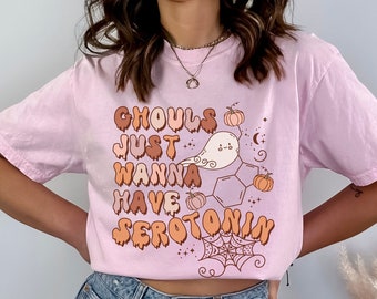 Puede incluir: Una camiseta rosa con un estampado gráfico que dice "Ghouls Just Wanna Have Serotonin" en una fuente que gotea. El gráfico también incluye un fantasma de dibujos animados, una telaraña y calabazas.
