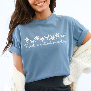 May include: Blue jean-colored t-shirt with the phrase "Practice radical empathy" in white script. The shirt features a floral and butterfly design above the text. The t-shirt is paired with black shorts.