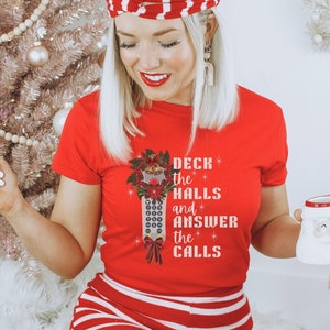 May include: A red t-shirt with a white and red striped design. The shirt features a graphic of a Christmas tree with a remote control and the text "Deck the Halls and Answer the Calls".