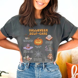 May include: A woman wearing a dark gray t-shirt with a graphic print that says "Halloween Self-Care" with various Halloween-themed illustrations. The shirt is a cropped style and the woman is wearing blue jeans.