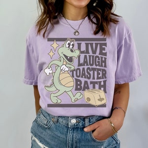 May include: A lilac-colored t-shirt with a cartoon dinosaur and toaster graphic. The text "LIVE LAUGH TOASTER BATH" is printed in a rectangular box. The shirt is a relaxed fit.