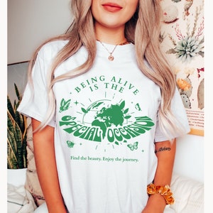 May include: White t-shirt with a green graphic of the Earth with butterflies and the text "Being Alive is the Special Occasion. Find the beauty. Enjoy the journey."