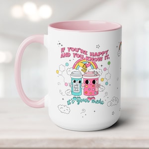 May include: White ceramic mug with a pink rim and handle. The mug features a colourful illustration of two pill bottles with faces and the text "If you're happy and you know it, it's your meds."