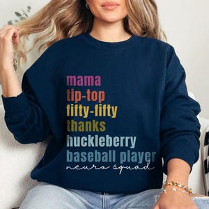 May include: Navy blue sweatshirt with colorful text: "mama, tip-top, fifty-fifty, thanks, huckleberry, baseball player, neuro squad." The word "NAVY" is at the bottom. The sweatshirt is worn by a person sitting on a white couch.