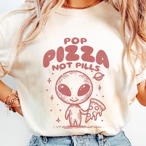 May include: A white t-shirt with a pink alien holding a pizza slice. The shirt has the text "Pop Pizza Not Pills" printed on it.