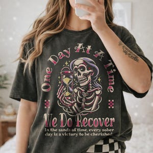 May include: Dark gray t-shirt with a graphic featuring a skeleton holding an hourglass, with text that reads "One Day At A Time" and "We Do Recover". The shirt has a vintage, worn look.