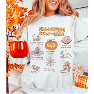 May include: A gray sweatshirt with a Halloween-themed design featuring illustrations of a pumpkin, a witch's hat, a spiderweb, and a tombstone. The text "Halloween Self-Care" is printed in orange and black letters.
