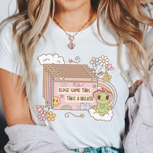 May include: White t-shirt with a pink and green graphic of a frog sitting on a cloud with the text "Close some tabs Take a breath".