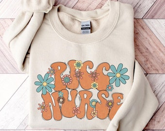 May include: A beige sweatshirt with the words "PUG NURSE" in orange and brown letters with flowers around the text. The sweatshirt is folded and laid on a wooden surface.