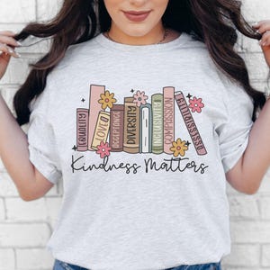 May include: A light gray t-shirt with a graphic of stacked books with words like "Equality", "Love", "Diversity", "Acceptance", "Compassion", and "Kindness Matters".
