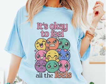 May include: Light blue t-shirt with the text "It's okay to feel all the feels" in pink and white. The shirt features a graphic of various emoji-like faces expressing different emotions. The word "CHAMBRAY" is printed at the bottom.