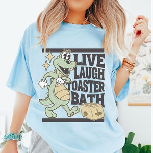 May include: Light blue t-shirt with the text "LIVE LAUGH TOASTER BATH" and a cartoon alligator with a toaster. The shirt is a chambray color.