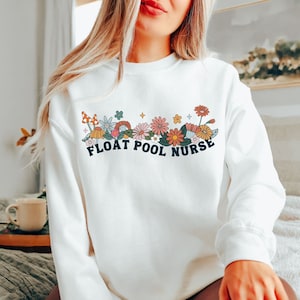 May include: White sweatshirt with a floral design and the text "FLOAT POOL NURSE" in a retro font. The design includes colorful flowers, mushrooms, and other botanical elements. The sweatshirt is a crew neck style.