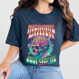 May include: A navy blue t-shirt with a graphic of a skull with a brain inside. The text "Migraine Brain Just Give Me Space!" is printed on the shirt.