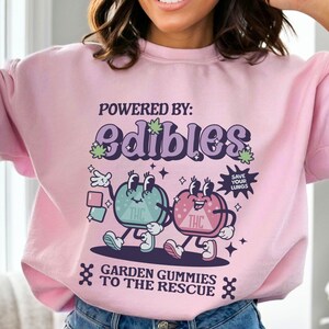 May include: A light pink sweatshirt with a graphic design featuring two cartoon gummies with the text "POWERED BY: edibles" and "GARDEN GUMMIES TO THE RESCUE".