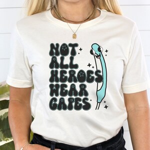 May include: A white t-shirt with a graphic print that says "Not All Heroes Wear Capes" in black with a blue and white cartoon character. The character is a stick figure with a large head and a blue body. The character is wearing a cape and has a star on its head.