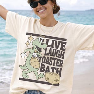 May include: Cream-colored t-shirt featuring a cartoon alligator and toaster with the words "LIVE LAUGH TOASTER BATH" in black. The alligator is light green with a beige belly and is walking. The toaster is beige.