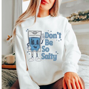 Op de afbeelding: Wit sweatshirt met een blauwe cartoon IV-zak met de tekst "Normal Saline" en de woorden "Don't Be So Salty" in blauw.