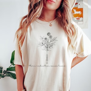 May include: A cream-coloured t-shirt with a black line drawing of a syringe with flowers growing from the needle. The text "harm reduction saves lives" is printed below the image.