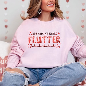May include: A pink sweatshirt with the text "You make my heart flutter" in red and white lettering. The text is surrounded by small red hearts. The sweatshirt is being worn by a person sitting on a couch.