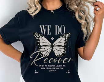 Pode incluir: Camiseta preta com um desenho de borboleta e as palavras "WE DO Recover". O design inclui o texto "WHEN WE RECOVER LOUDLY, WE KEEP OTHERS FROM DYING QUIETLY". Uma camiseta de estilo casual e confortável.