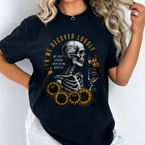 May include: Black t-shirt with a graphic of a skeleton holding a butterfly and sunflowers. The text on the shirt reads "When we recover loudly we keep others from dying quietly."