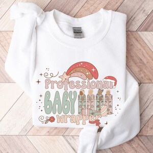 May include: White sweatshirt featuring the text "Professional Baby Wrapper" in a retro font, with a rainbow and star graphic. The design includes wrapped gifts and a Santa hat, ideal for the festive season.